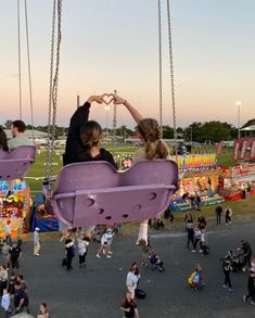 two people sitting on swings in the middle of an amusement park while others walk around