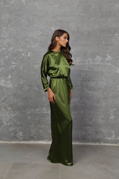 a woman in a long green dress stands against a gray wall and looks off to the side