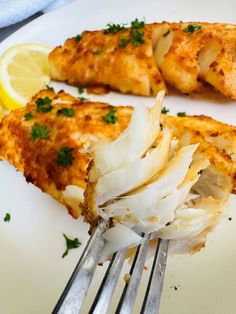 two pieces of fish on a white plate with a fork and lemon wedges next to it