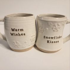 two coffee mugs with words on them sitting side by side, one has snowflakes and the other is saying warm wishes