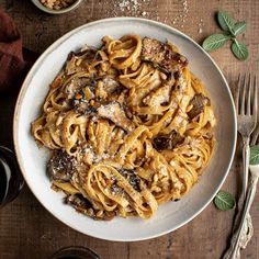 a white plate topped with pasta covered in sauce and mushrooms next to silverware on a wooden table