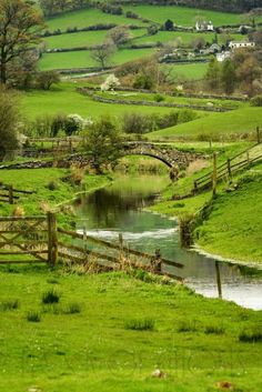 a small stream running through a lush green field next to a wooden fence and stone bridge