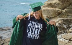 a woman in a graduation cap and gown is standing on the rocks by the water