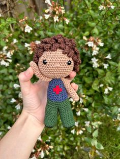 a hand holding a small crocheted doll in front of some bushes and flowers