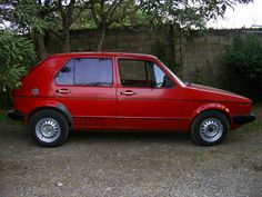 a small red car parked in front of a tree and shrubbery behind the vehicle