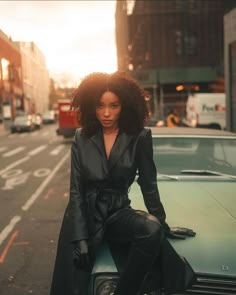 a woman is sitting on the hood of a car in an urban setting, wearing all black