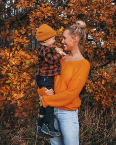 a woman in an orange sweater holding a small child while standing next to trees with yellow leaves