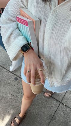 a woman holding an open book and a drink in her hand while standing on the sidewalk