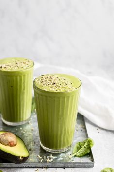 two glasses filled with green smoothie next to an avocado on a tray