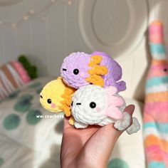 a hand holding three small crocheted toys in front of a bed with pillows