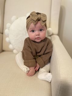 a baby sitting on a couch wearing a brown sweater with a bow in it's hair