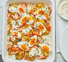 a white dish filled with eggs and salmon on top of potato wedges next to a bowl of ranch dressing