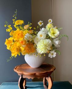 a white vase filled with yellow and white flowers on top of a wooden table next to a blue wall