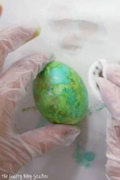 two hands in white gloves are holding an egg with green paint on it and another hand is wearing pink gloves