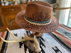 Rustic western rancher features a hand-burned floral pattern embellished with turquoise stones. The custom band includes leather, lace, feathers, and glass beads. Add a touch of Southwestern charm to any Fall outfit. One size fits most and inside band is adjustable. Fitted Rustic Hat Bands For Festivals, Bohemian Hat Bands With Feathers For Ranch, Bohemian Hat Band With Feathers For Ranch, Rustic Distressed Brown Hat Band For Ranch, Bohemian Concho Hat For Country Events, Bohemian Concho Hat Bands For Country Events, Bohemian Feather Hat Bands For Ranch, Adjustable Southwestern Hat Band For Western-themed Events, Western Hat Bands With Leather Sweatband For Western-themed Events