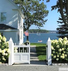 a white picket fence in front of a house with flowers and trees on the side