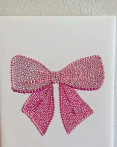 the pink bow is made out of small beads on white paper and hangs on a wall