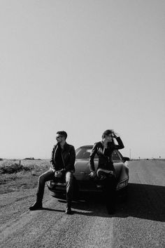 two people sitting on the hood of a car in the middle of an empty road