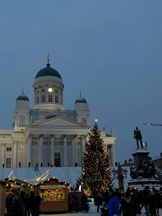 a large white building with a christmas tree in the foreground and people walking around it