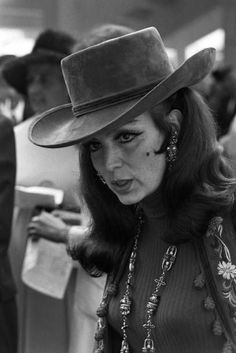 a woman wearing a hat and necklaces standing in front of a man with a suit on