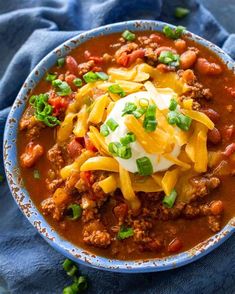 a bowl filled with chili and cheese on top of a blue cloth next to a spoon