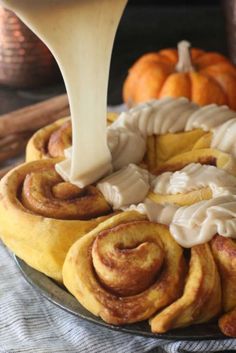 a cinnamon roll being drizzled with icing on a plate next to pumpkins