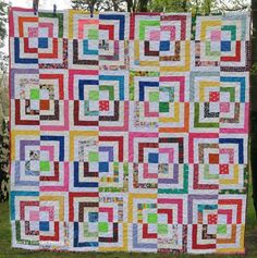 a multicolored quilt hanging from a tree in the grass with trees in the background