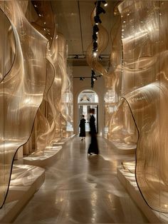 two people walking down a long hallway lined with gold colored walls and suspended lights on the ceiling