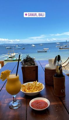 there is a plate of fries and a drink on the table near the water with boats in the background