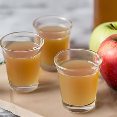 three shot glasses filled with liquid next to an apple on a cutting board in front of two apples