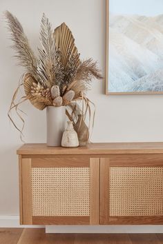 a vase filled with dried plants sitting on top of a wooden cabinet next to a painting