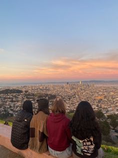 three people are sitting on a ledge looking at the city from atop a hill in front of them