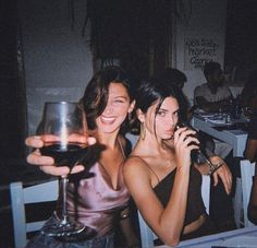 two women sitting at a table with wine glasses in front of them and one holding a cell phone to her ear