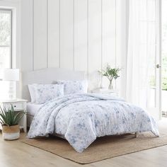 a white bed with blue flowers on it in a room next to a window and potted plant