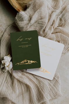 two green and gold wedding cards on top of a white blanket next to a flower