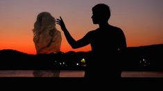 two people standing next to each other in front of a body of water at sunset