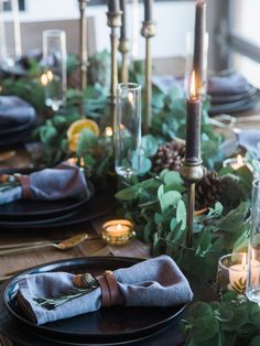 the table is set with black plates and silverware, greenery, candles, and napkins