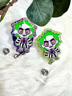 Beetlejuice-Inspired Badge Reels - Spooky & Fun! Add a touch of spook-tacular charm to your workday with these Beetlejuice-inspired badge reels! Featuring an adorable yet mischievous rendition of the popular character, these badge reels are perfect for fans of the classic film. Each reel showcases a mini Beetlejuice with his signature wild green hair, pale face, and striped suit, brought to life in a vibrant and eye-catching design. The reels are accented with either a purple or green glitter border, adding an extra pop of fun. These badge reels are both functional and stylish, with a retractable cord and a clip that easily attaches to your ID or keycard. Whether you're dressing up for Halloween or just want to show off your love for all things creepy, these Beetlejuice badge reels are a m Themed Multicolor Badge Holders As Gifts, Themed Multicolor Badge Holders For Gift, Fun Black Badge Reel For Gifts, Customizable Novelty Multicolor Badge Reel, Customizable Multicolor Novelty Badge Reel, Beetlejuice Characters, Nurse Accessories, Pale Face, Striped Suit