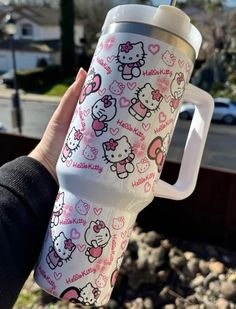 someone holding up a hello kitty travel mug in their left hand, with hearts and bows on it