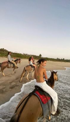 three people riding horses on the beach with other people in the water and one person walking behind them