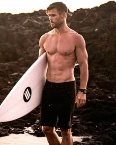 a shirtless man holding a white surfboard on the beach in front of rocks