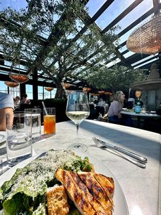 a white plate topped with meat and salad next to glasses of wine on top of a table