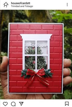 a hand holding up a red card with a window and christmas decorations on the windowsill