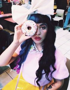 a woman with blue hair holding a doughnut up to her eye and wearing a white bow