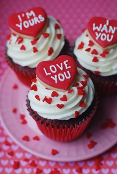 three cupcakes with white frosting and red heart decorations on a pink plate