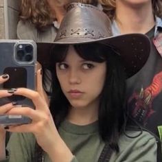 a woman wearing a cowboy hat taking a selfie with her cell phone while standing in front of other people