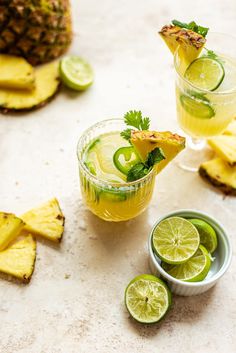 two glasses filled with drinks next to sliced pineapples and lime slices on a table