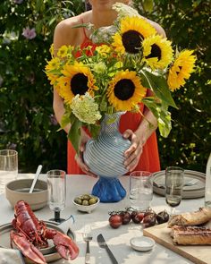 a woman holding a vase with sunflowers in it on top of a table