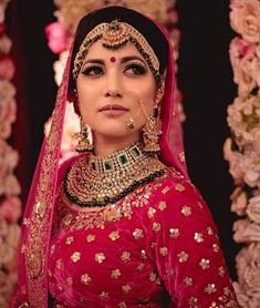 a woman wearing a red and gold bridal outfit with jewelry on her head, in front of flowers