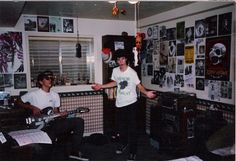 two young men are playing guitars in a room with posters on the wall and floor